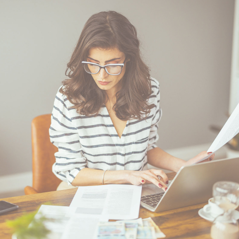 Picture of a woman sitting at a desk while working on her laptop and looking at various files 